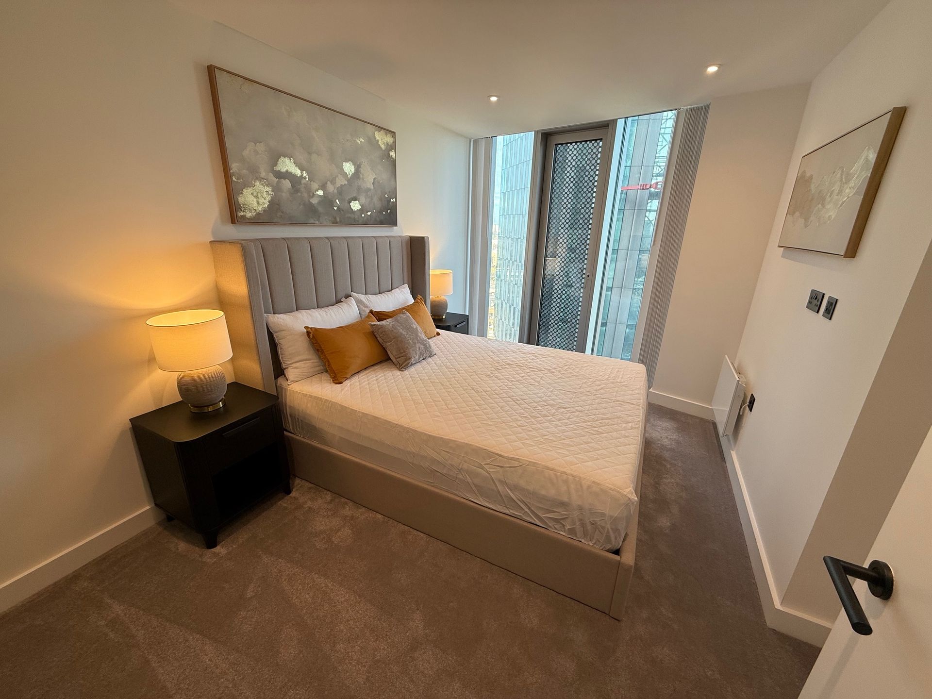 Bedroom with a bed, nightstand, lamps, and a large window with a city view.