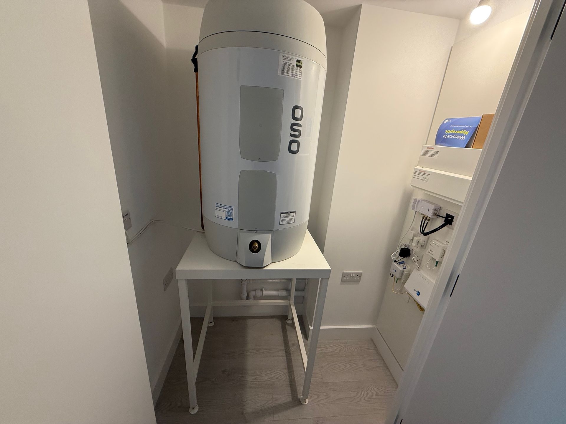 White hot water heater on a white table in a small room. The room has light grey walls and a white door.