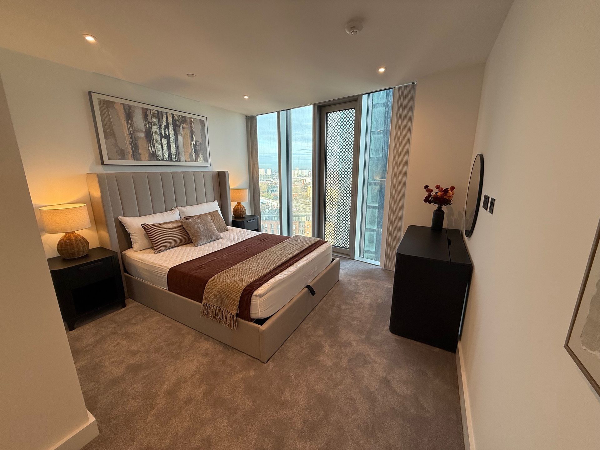 Bedroom with bed, nightstands, dresser, large window. Soft carpet, neutral colors, modern decor.