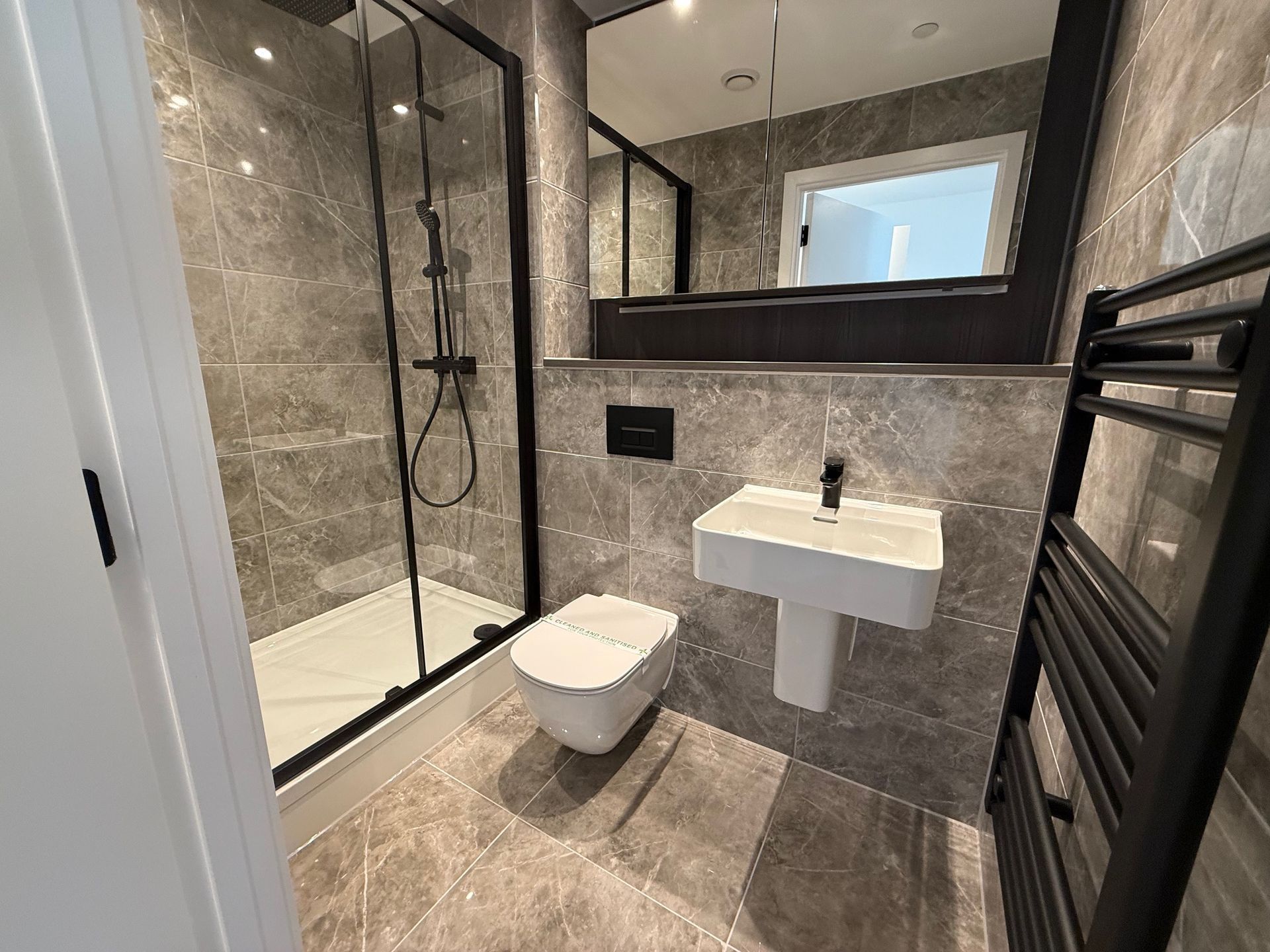 Modern bathroom with gray tiles, a black shower frame, toilet, sink, and towel warmer.