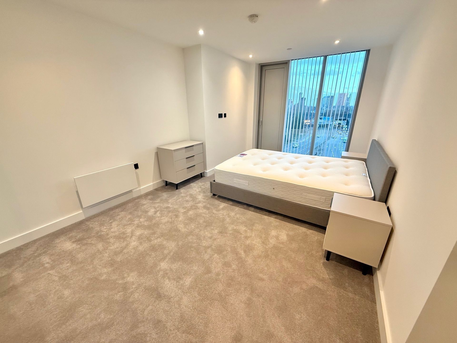Bedroom with bed, nightstands, dresser, large window. Beige carpet and white walls.