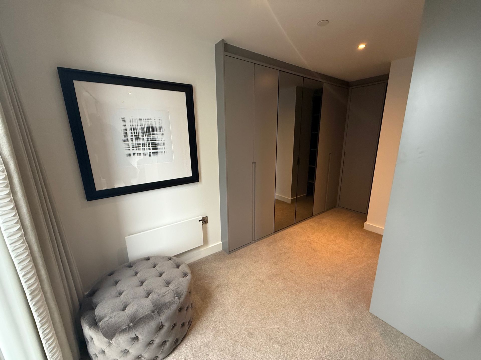 Walk-in closet with gray doors, mirror, art, and a tufted ottoman. Neutral carpet and walls.