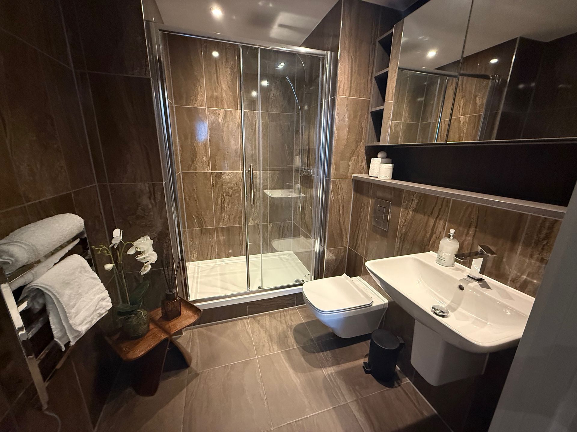 Brown tiled bathroom with glass shower, floating toilet, sink, mirror, and towel rack.