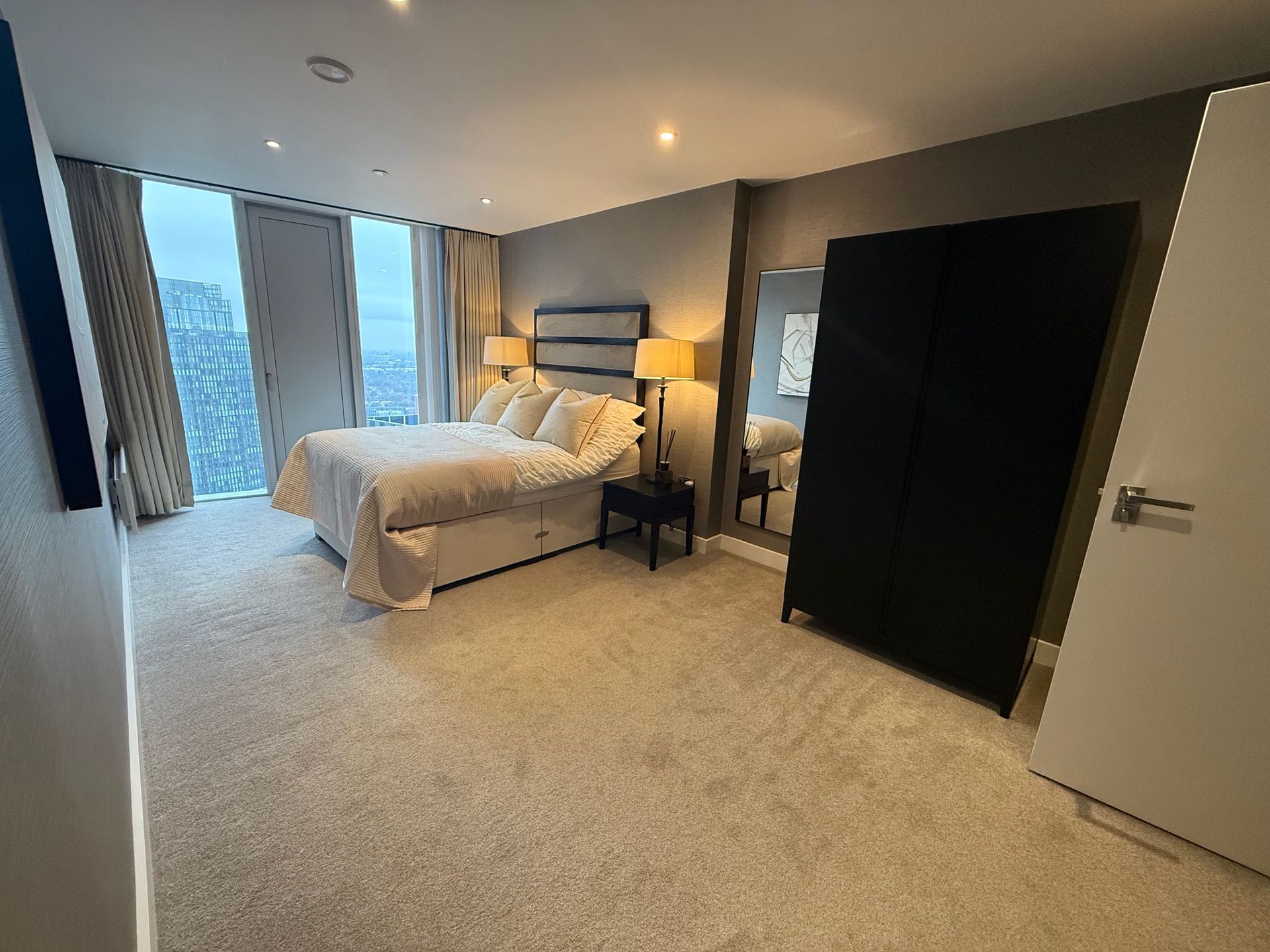 Bedroom with bed, closet, mirror, and large windows with a city view.