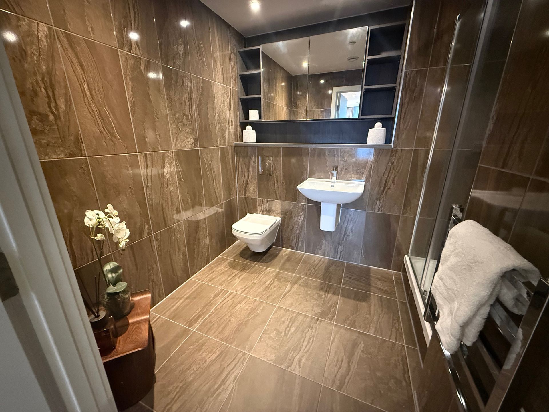 A small, modern bathroom with brown tile walls and floor. A toilet, sink, and mirror are visible. A towel hangs on a radiator.