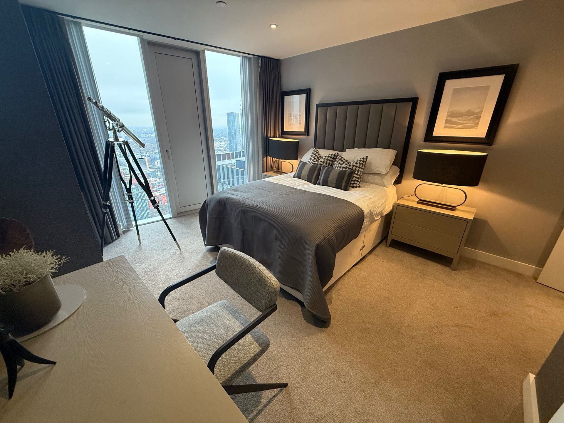 Bedroom with bed, desk, and large window overlooking a cityscape. Gray and neutral tones.