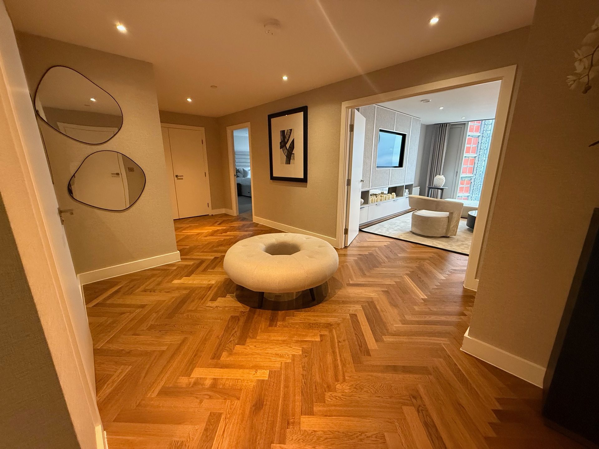 Hallway with herringbone wood floors, a round ottoman, mirrors, and an open doorway to a living room.