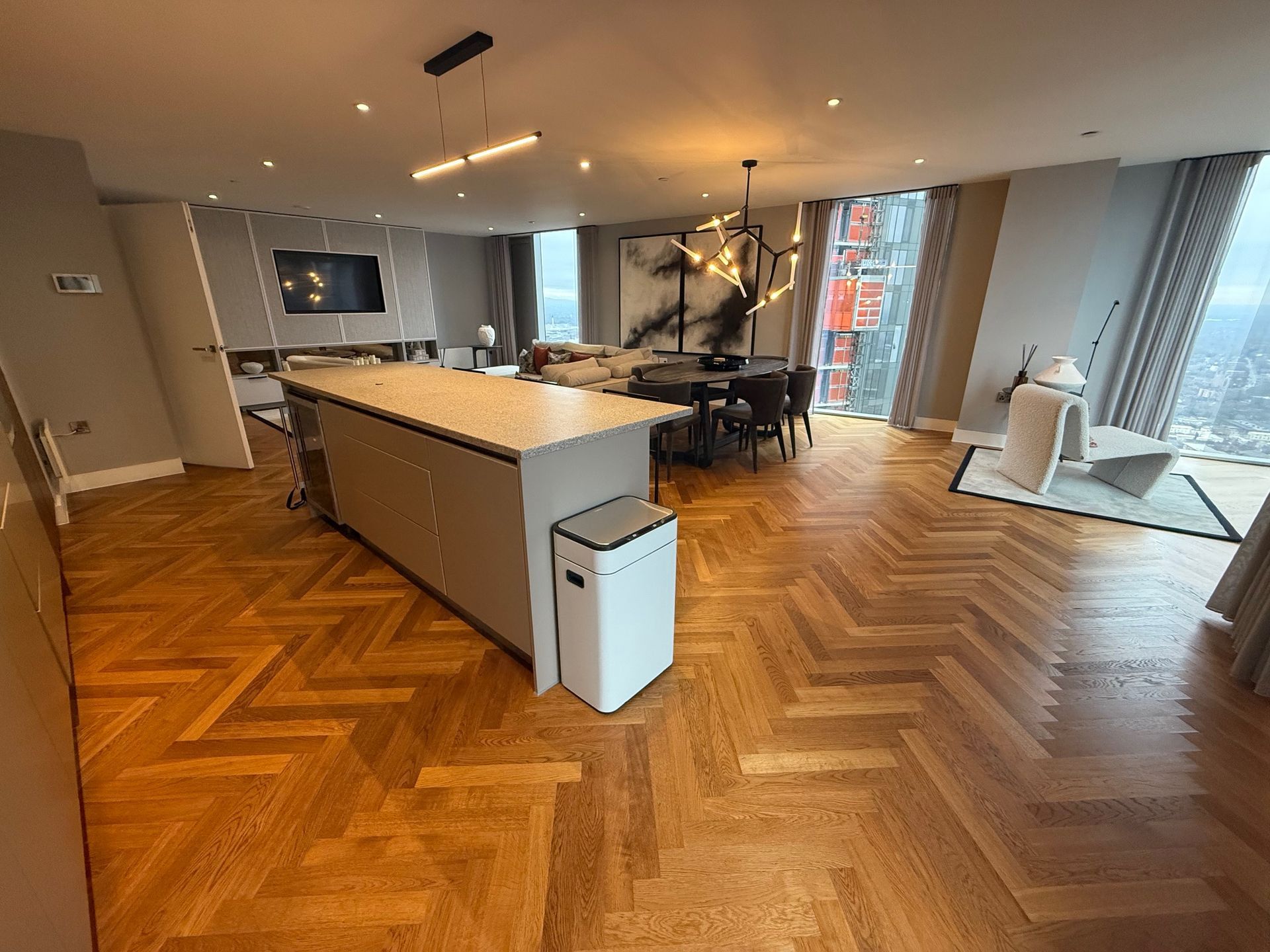 Open-plan luxury apartment with wood parquet floor, kitchen island, dining area, and city views through large windows.