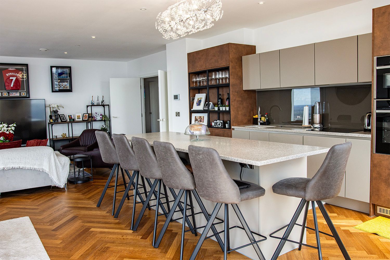 Modern kitchen with island seating and open living space. Herringbone floors, neutral tones, and a bedroom visible.