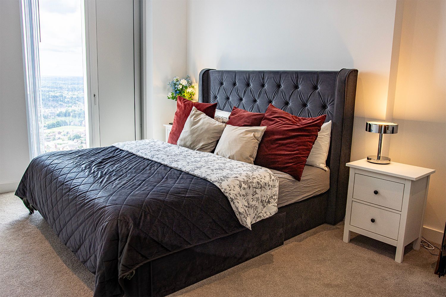 Bedroom with a gray upholstered bed, decorative pillows, white nightstand, and window.