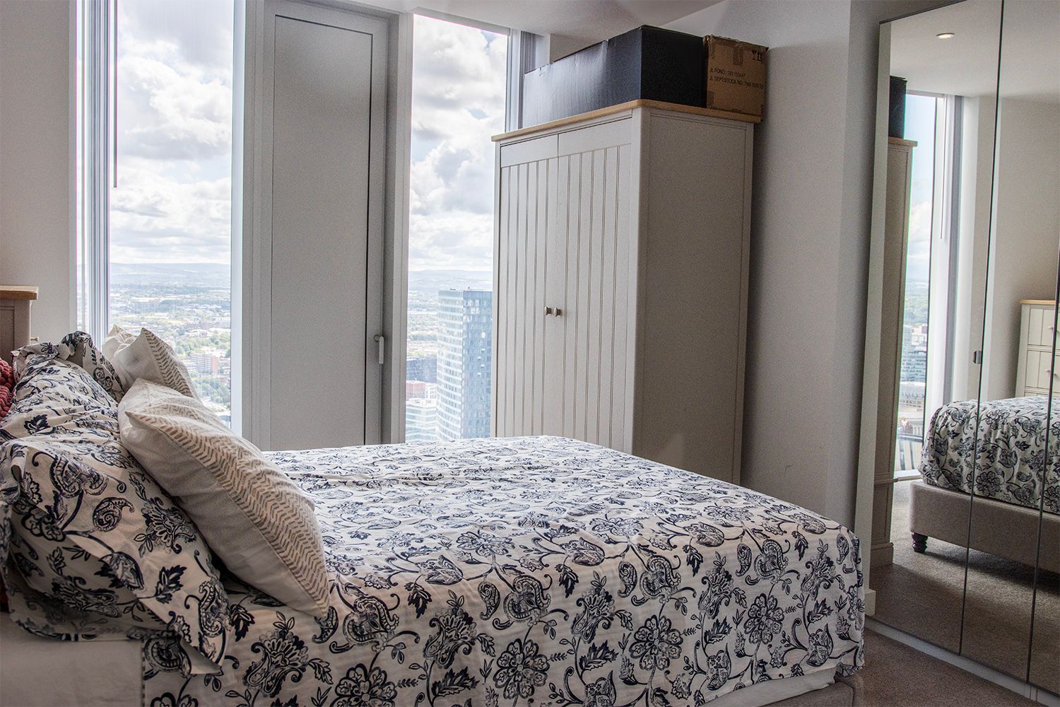 Bedroom with bed, white wardrobe, large window with city view, and mirrored closet door.