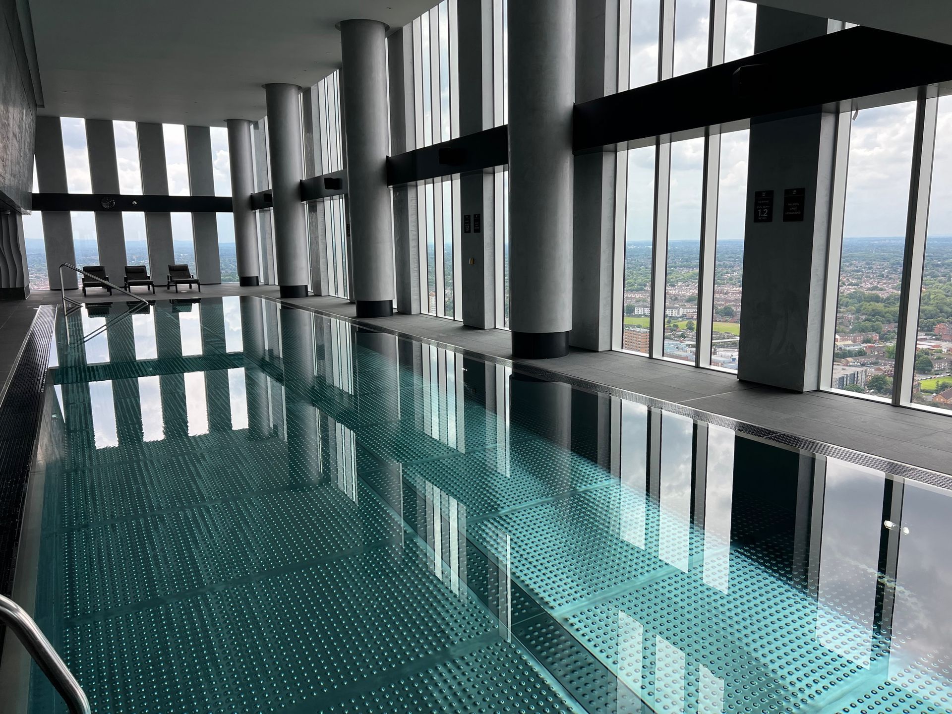 Indoor swimming pool with city view through large windows. Reflective water, gray pillars, and a distant cityscape.