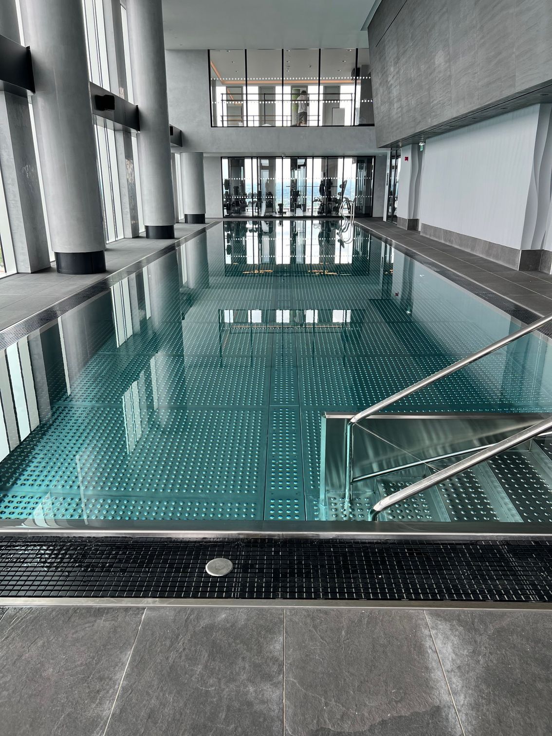Indoor swimming pool with turquoise water, surrounded by glass windows and gray columns.