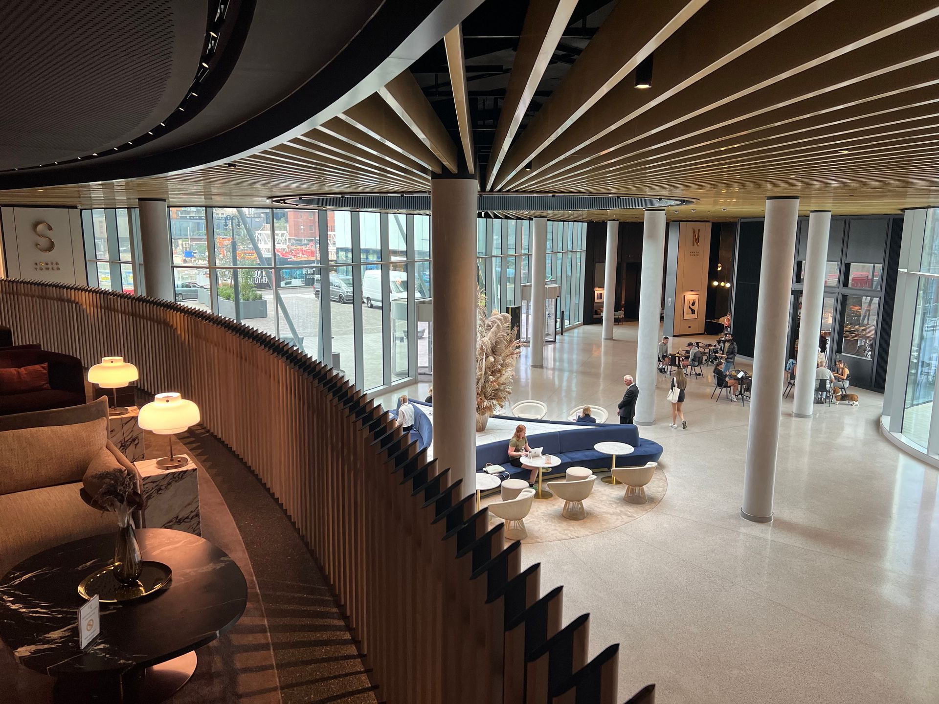 Interior view: curved balcony with seating overlooks a modern lobby with columns, seating, and a glass exterior.
