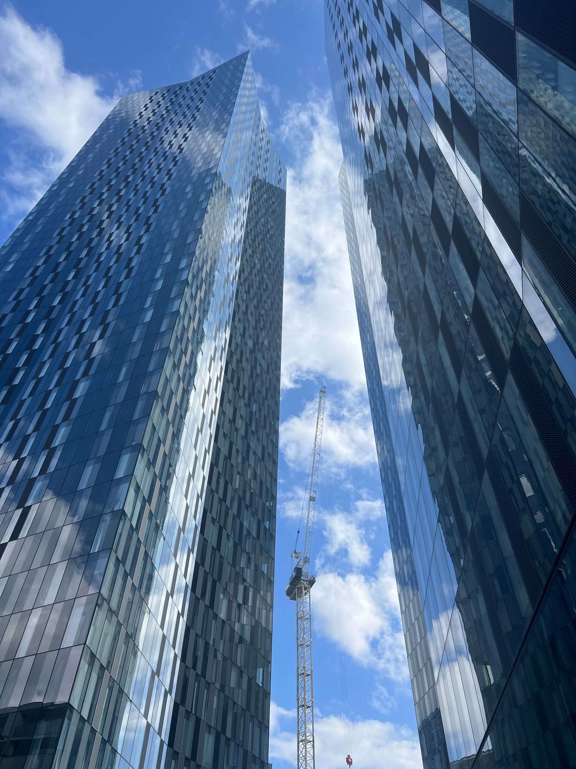 Two tall glass skyscrapers reaching towards a blue sky with clouds; a construction crane in between.