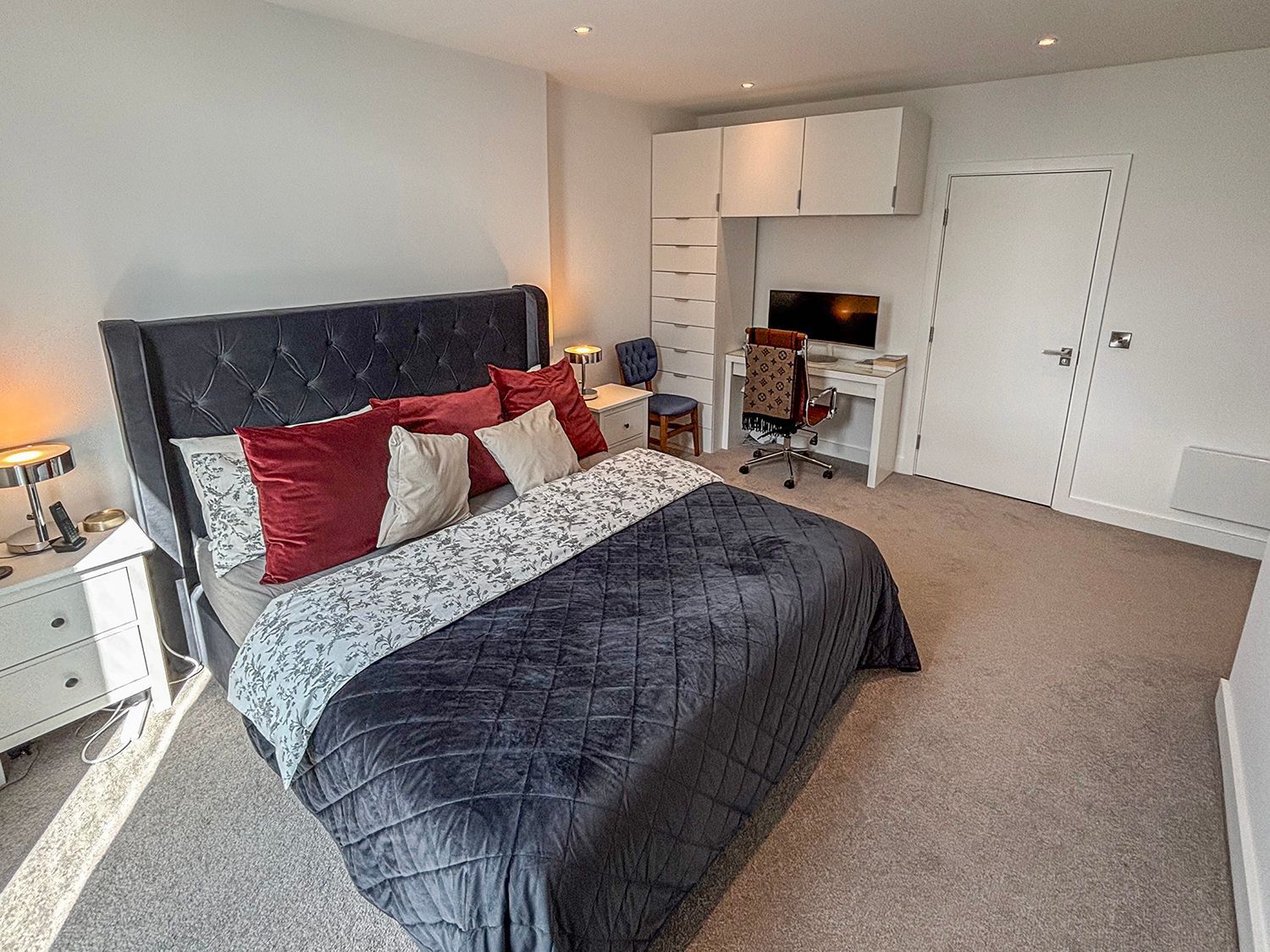 Bedroom with bed, desk, and built-in storage. Soft carpet, white walls, and a closed door.