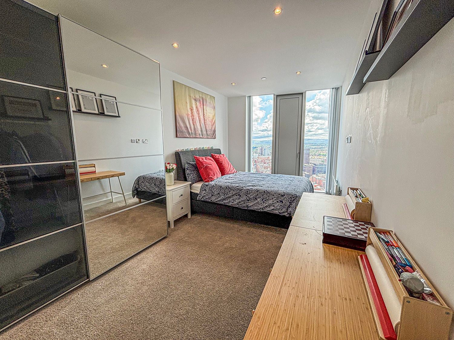 Bedroom with bed, mirrored wardrobe, desk, and city view from the window.