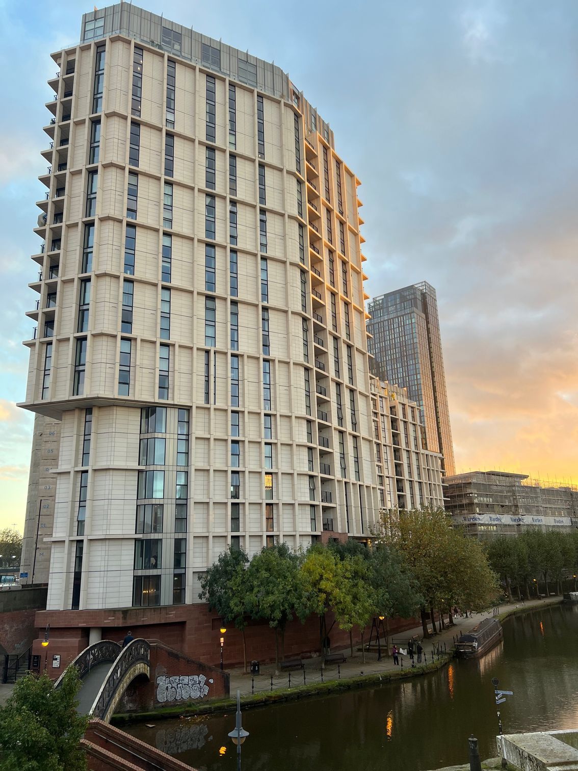 Two tall apartment buildings next to a canal with a bridge, at sunset.