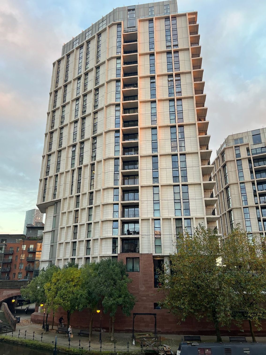 Tall, light-colored apartment building with many windows, next to a canal. Trees and other buildings are visible.