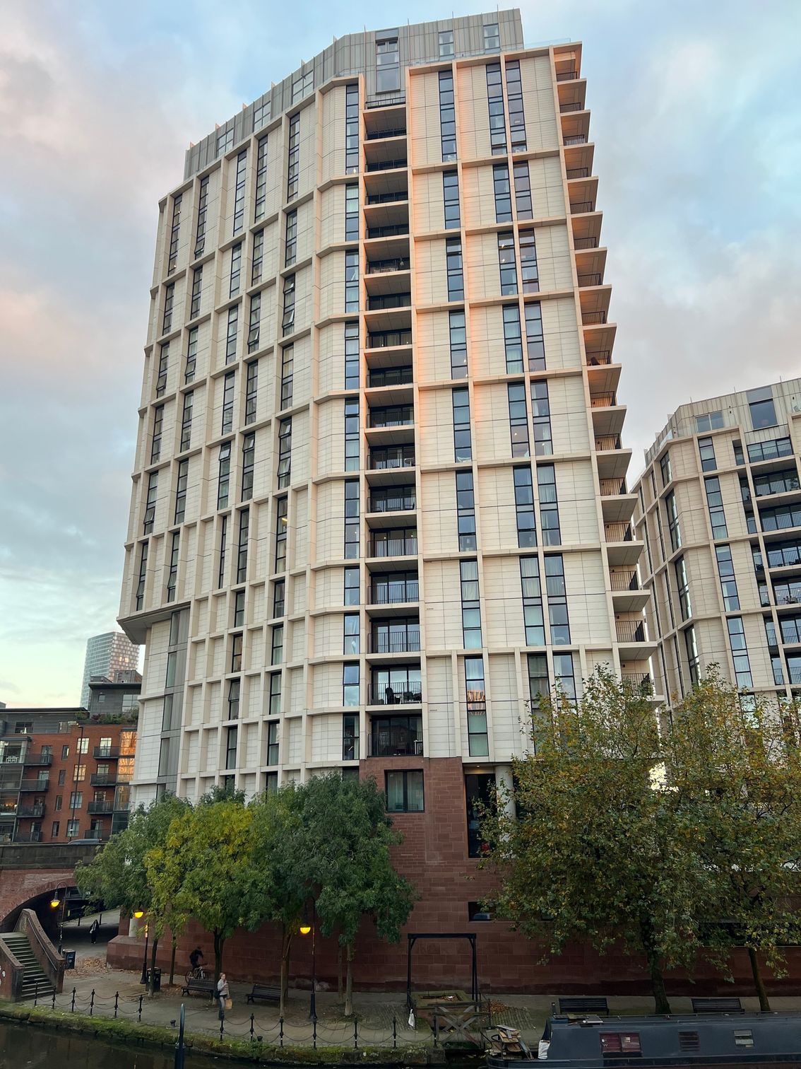 Tall modern apartment building with balconies, near a canal and trees.