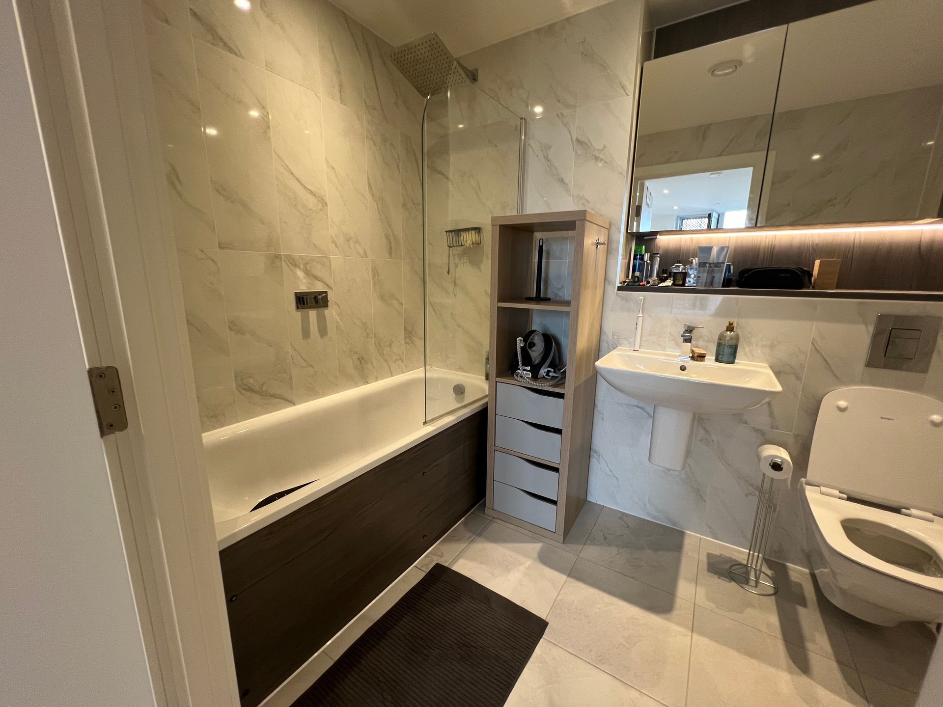 Bathroom with marble-effect walls, a bathtub, storage unit, sink, toilet, and mirror.