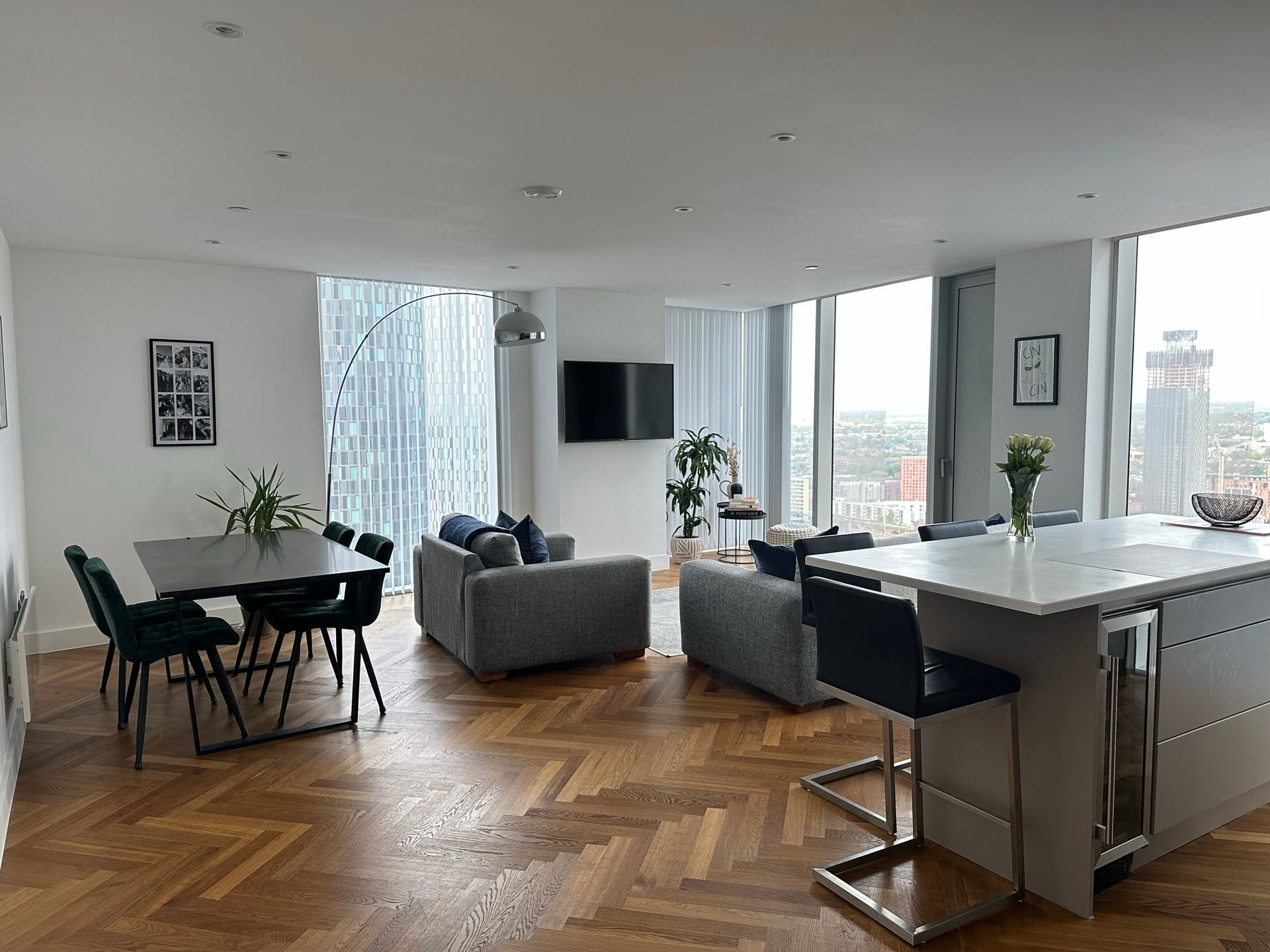 Open-plan living space with a dining area, two gray sofas, kitchen island, and large windows with city views.