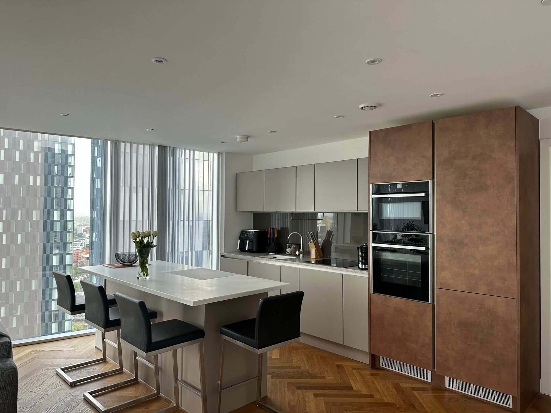 Modern kitchen with island, bar stools, and city view through a large window. Brown and grey cabinetry.