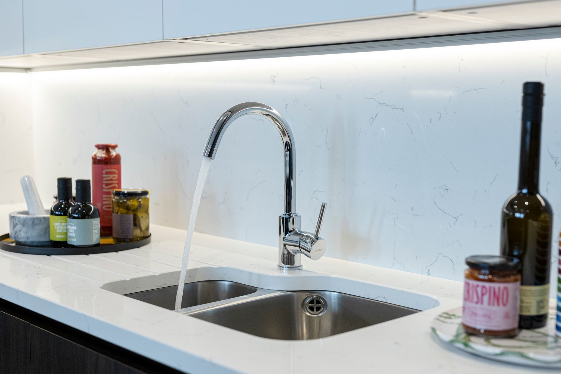 Kitchen sink with running water, countertop with jars and bottles, under cabinet lighting.