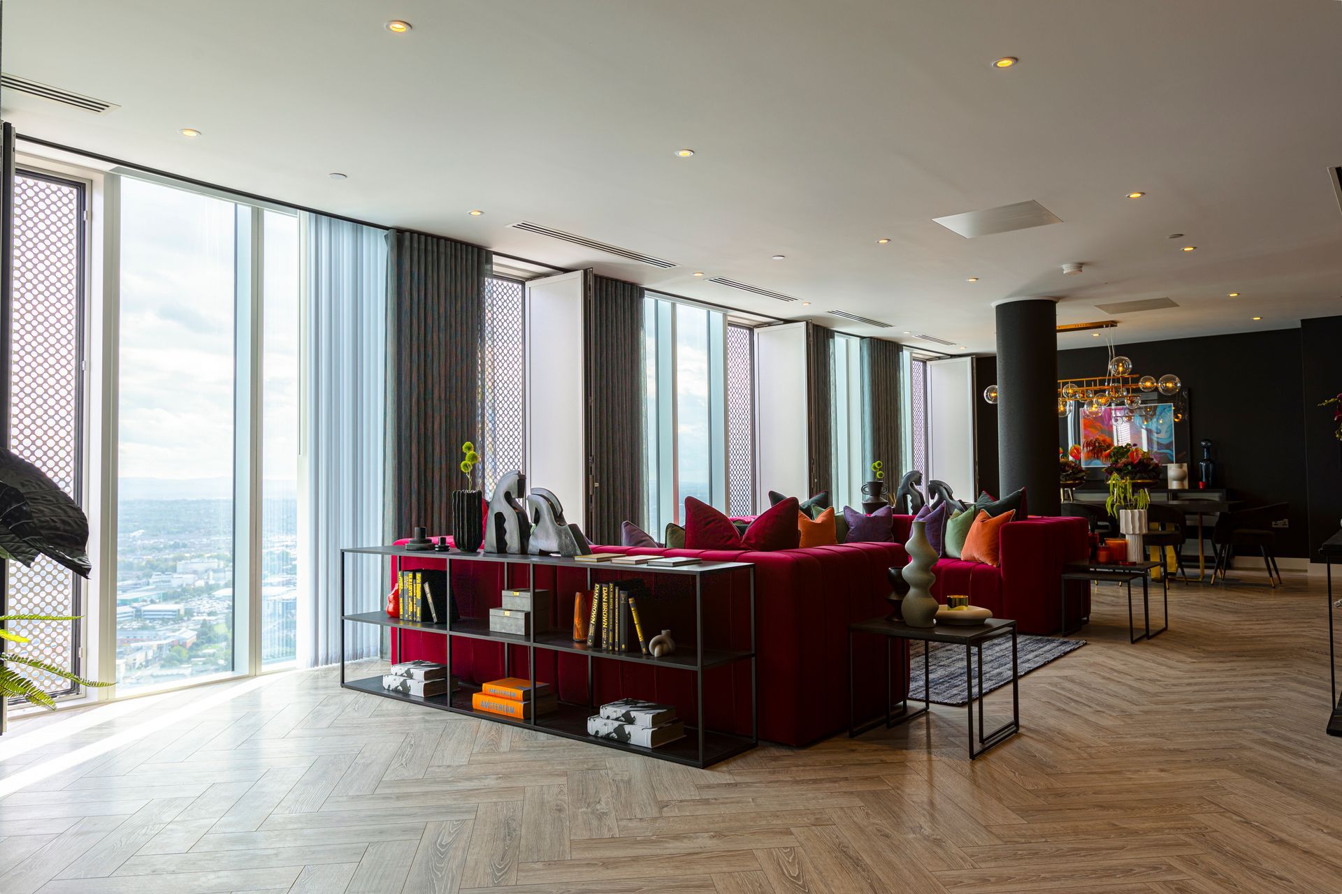 Living room with red sofa, floor-to-ceiling windows, and city view. Neutral tones with colorful accents.