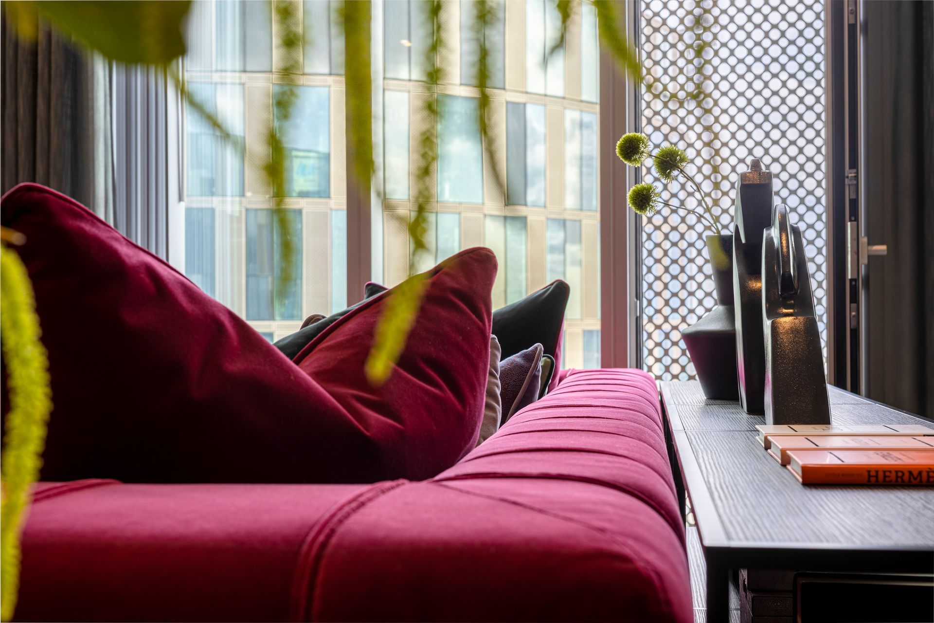 Red velvet sofa with pillows near a window with cityscape views and decorative objects.