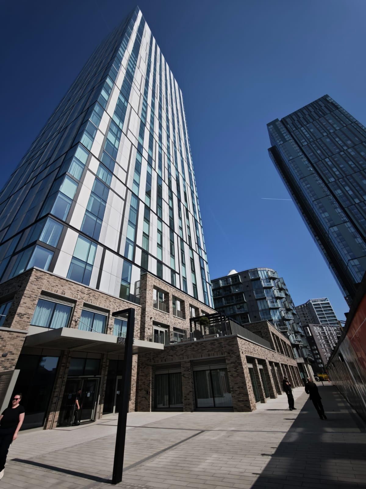 Two tall modern buildings under a blue sky, one with brick base, walkway and people.