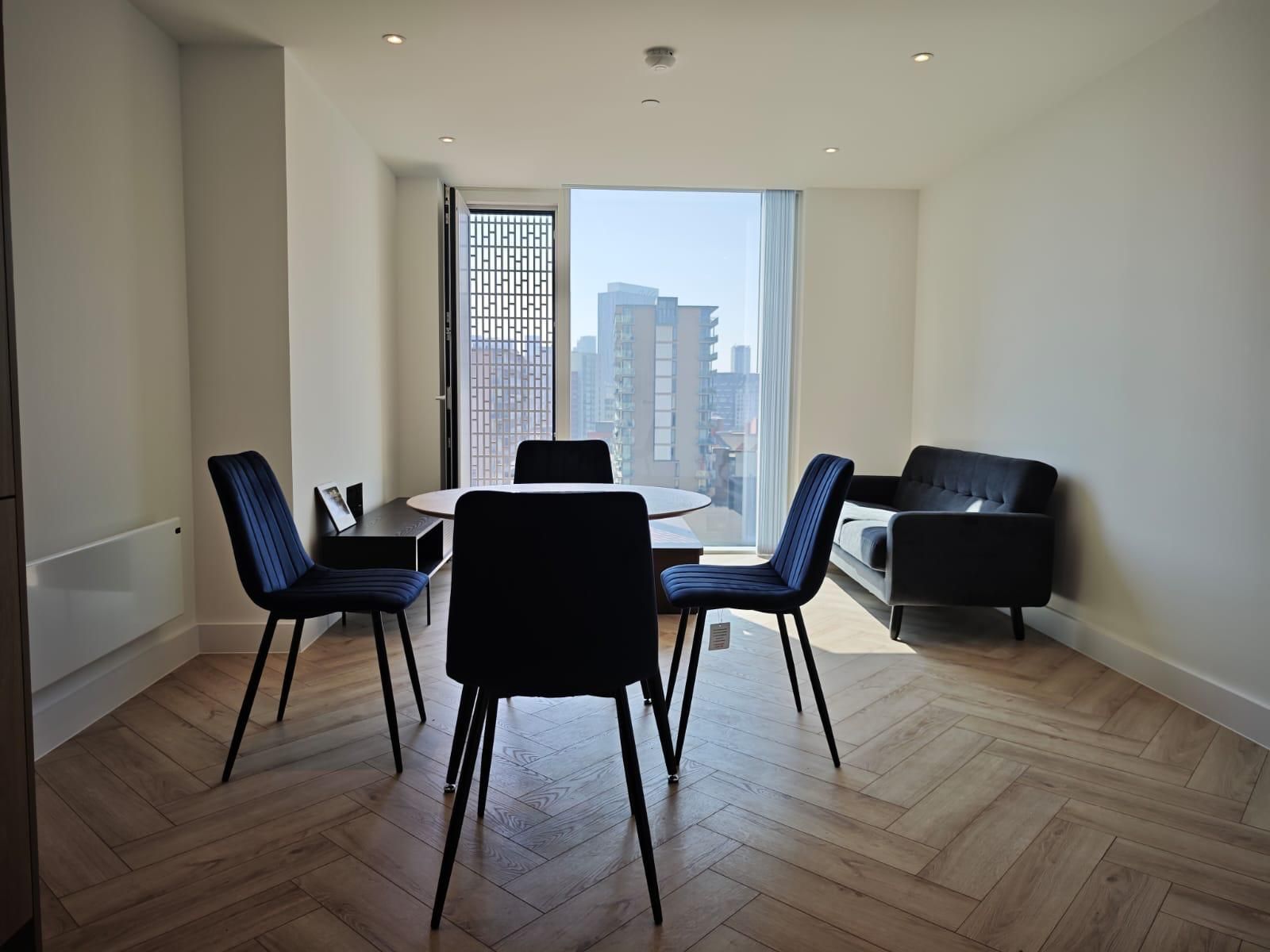 Dining area with round table, four blue chairs, small black sofa, and city view through large window.