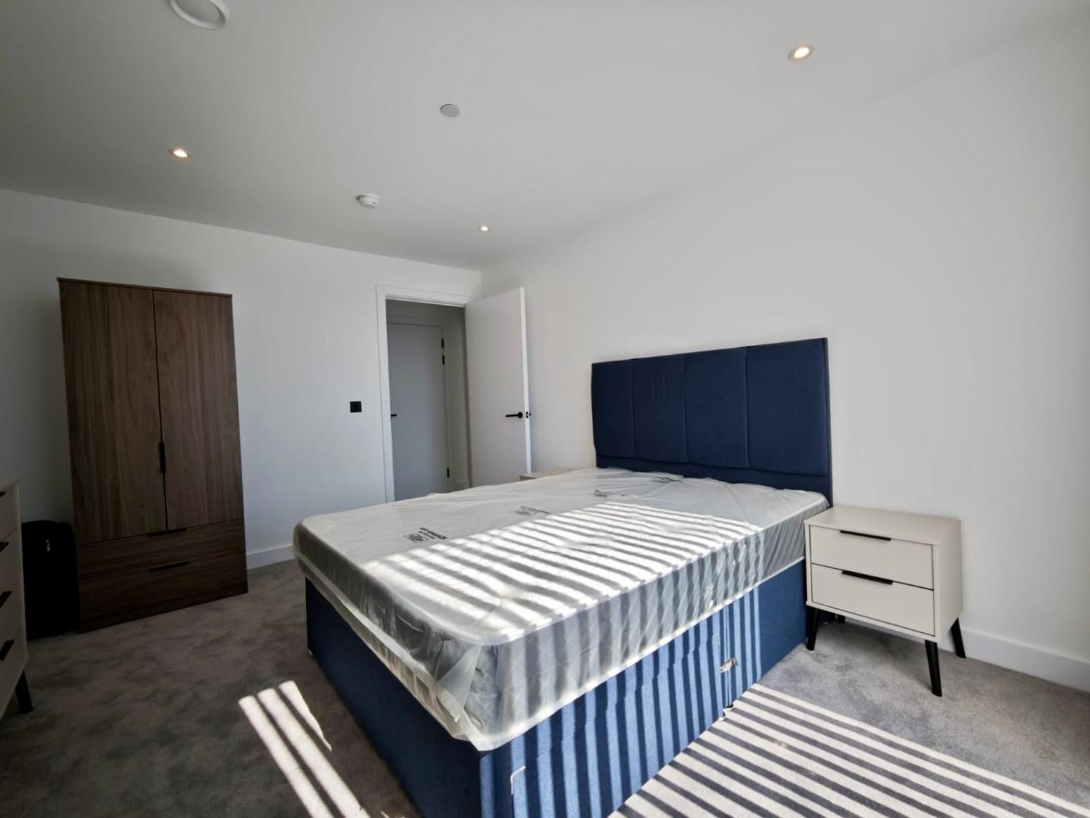 Bedroom with a blue bed, nightstand, and wardrobe on a carpeted floor. White walls.