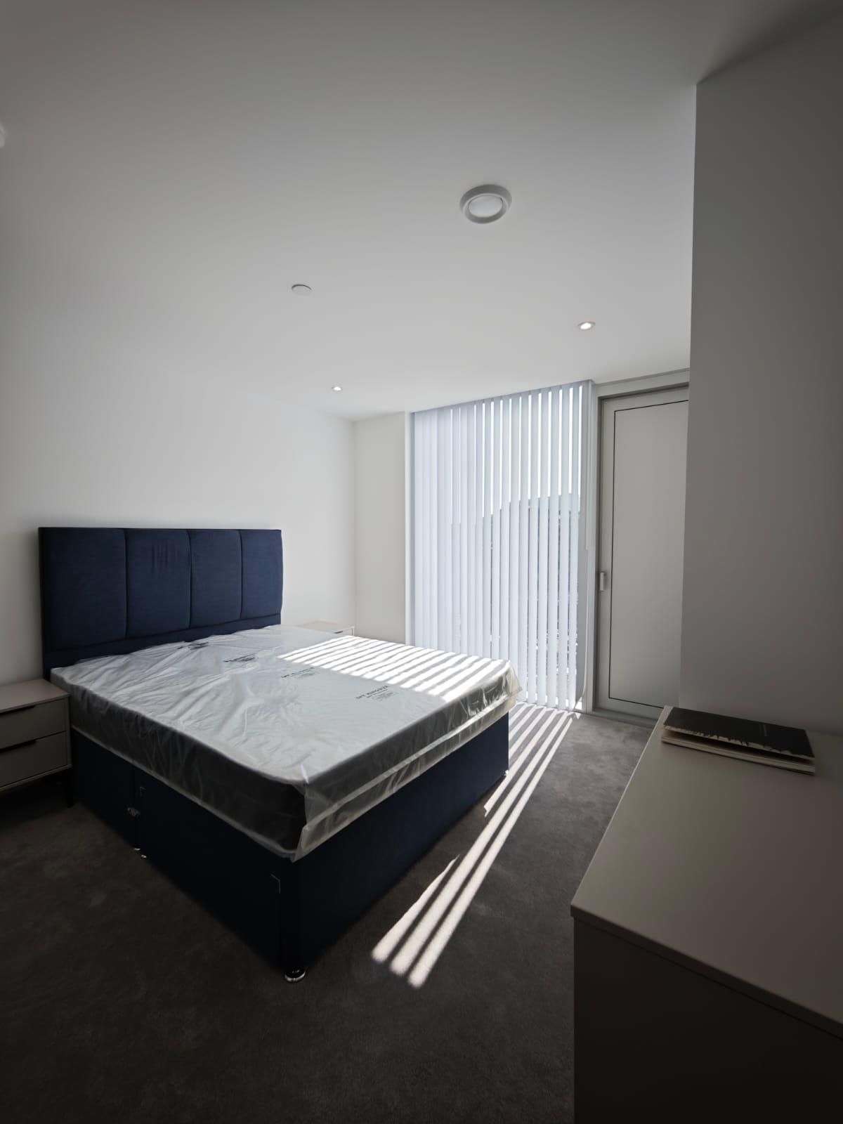 Bedroom with a blue bed, nightstand, and desk. Sunlight streams through the sheer curtains on the window.