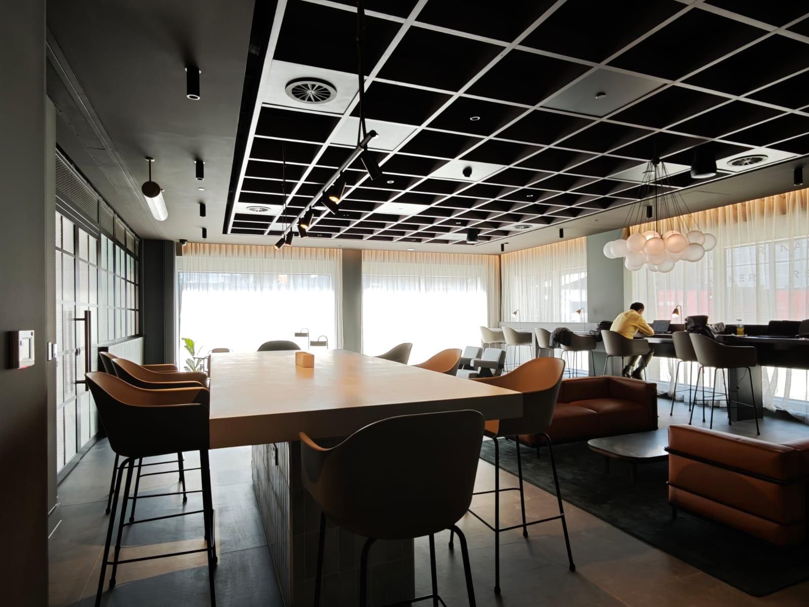 Modern office lounge with large table and chairs. Black grid ceiling, large windows, and a person walking in the background.