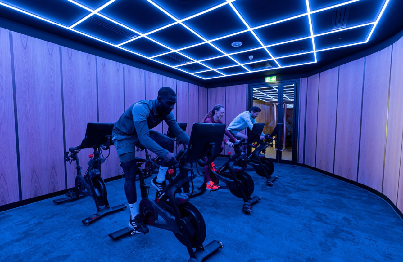 Three people cycling in a room with blue neon ceiling lights and wood-paneled walls.