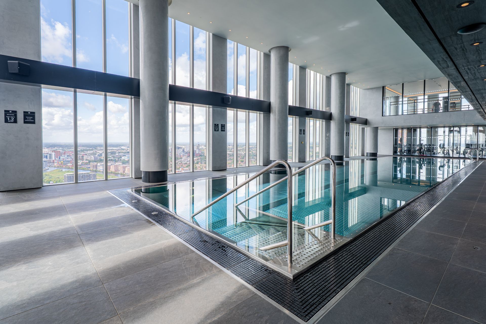 Indoor swimming pool with city view, large windows, pillars, and stainless steel handrails.