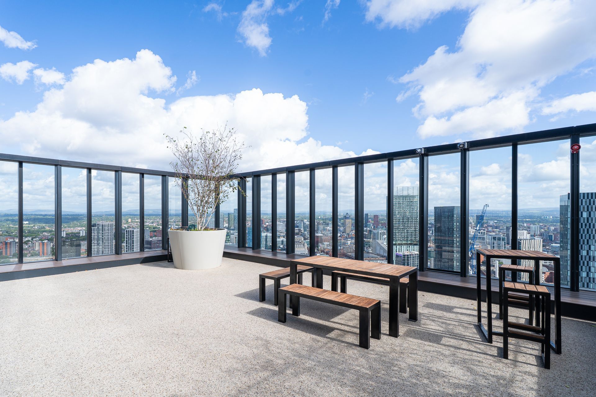 Rooftop terrace with tables, benches, and a large plant pot, offering city views under a blue sky with clouds.