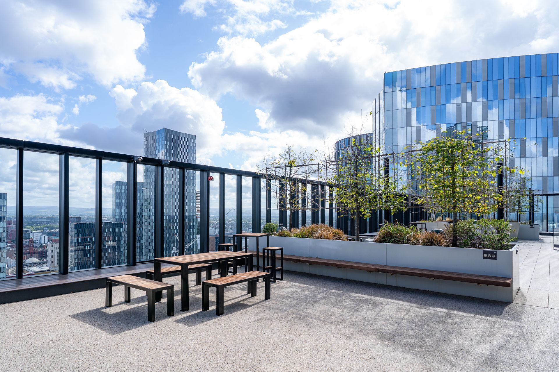 Rooftop terrace with seating and plants, overlooking a cityscape under a partly cloudy sky.