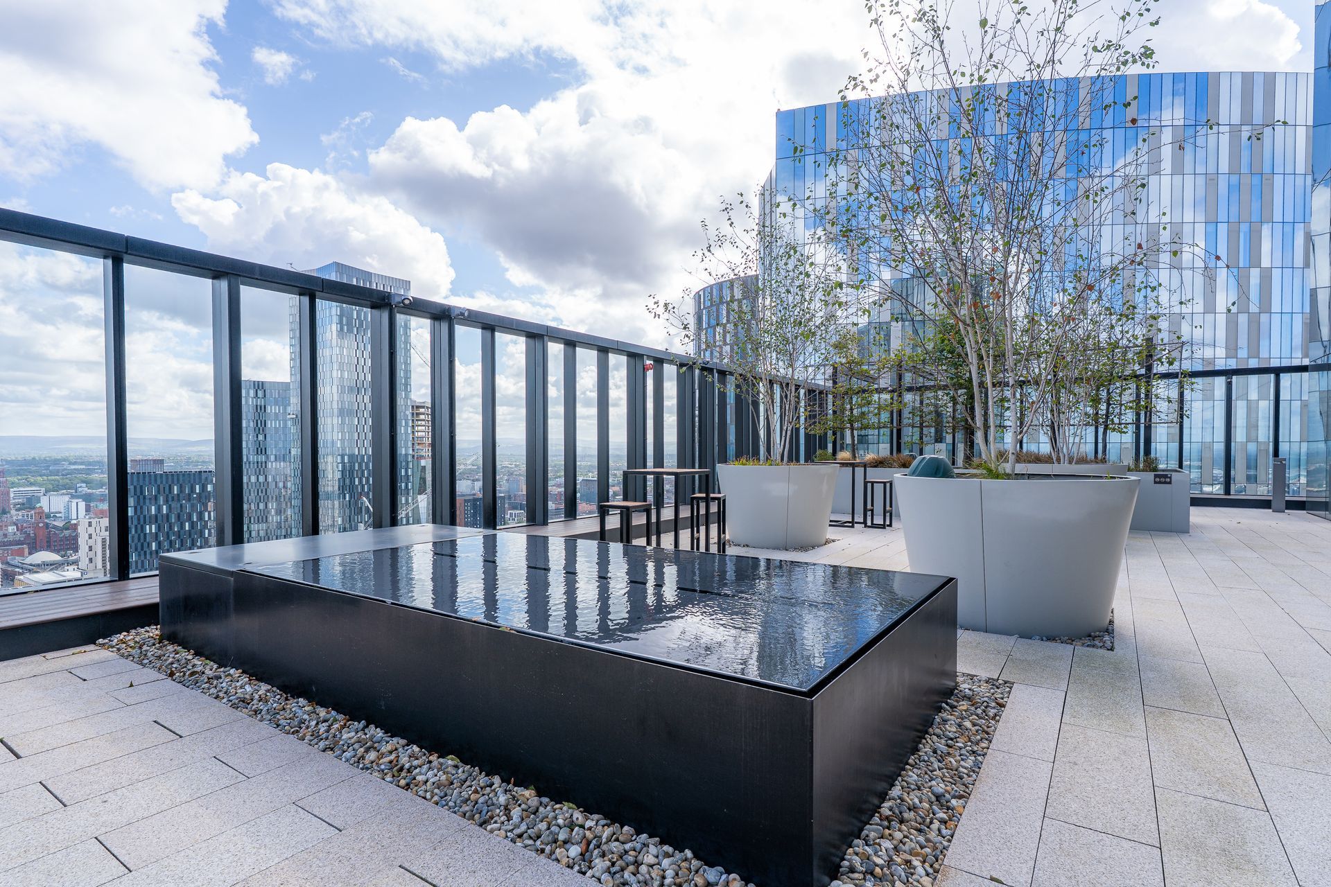 Rooftop patio with a reflecting pool, potted trees, and city view, set against a cloudy sky.
