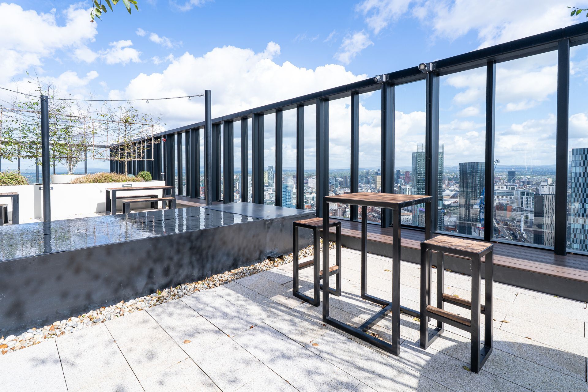 Rooftop patio with high-top table and stools, overlooking a city. Metal railing and blue sky.