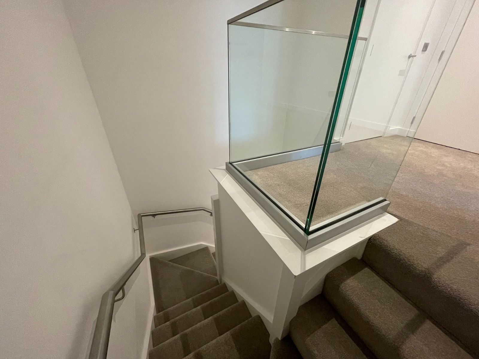 Staircase with carpeted steps leading down. Glass railing on a landing with white walls and a metal handrail.