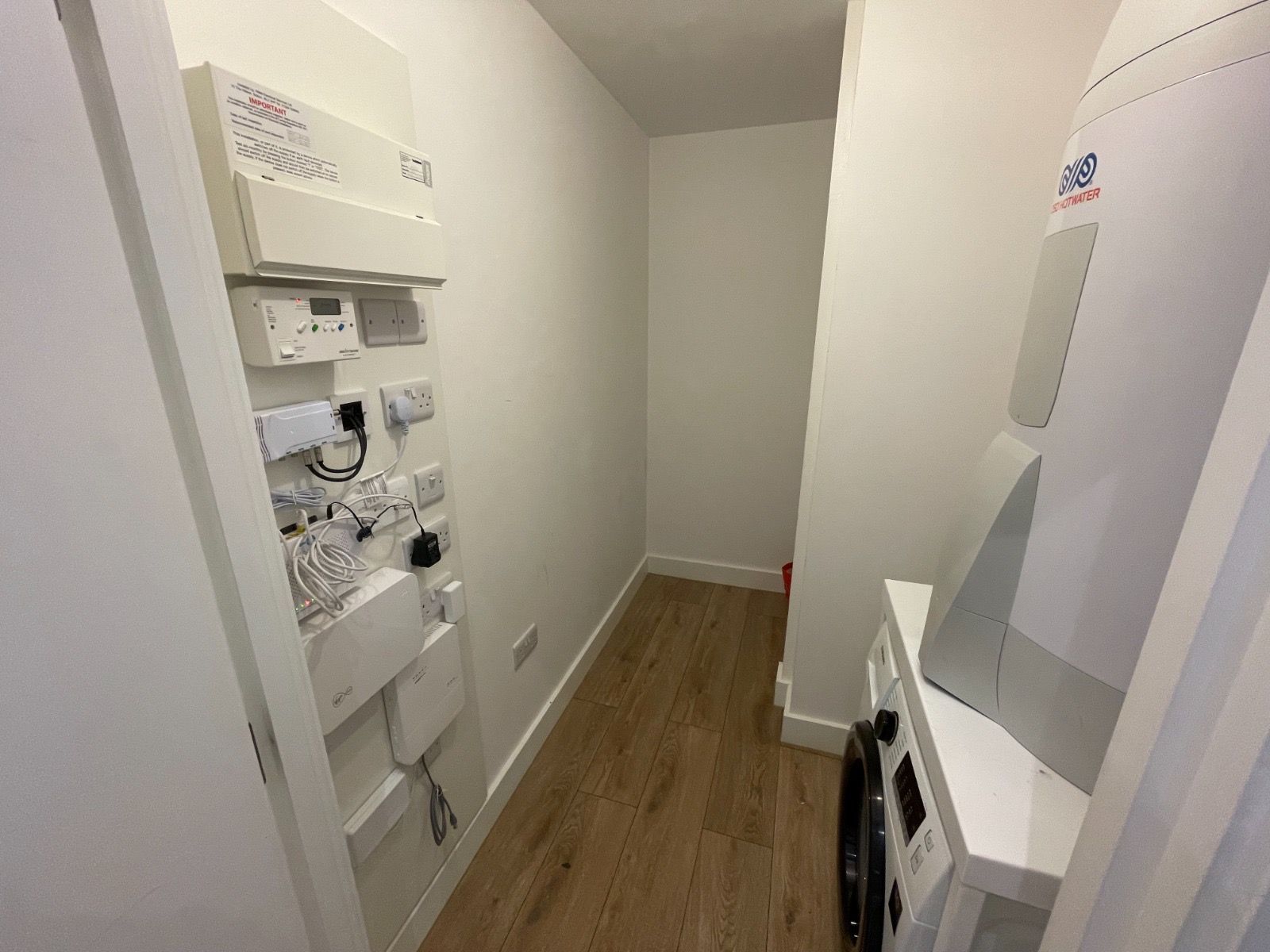 Narrow utility room with white walls, wood floor, and electrical panels. Laundry machine is on the right.