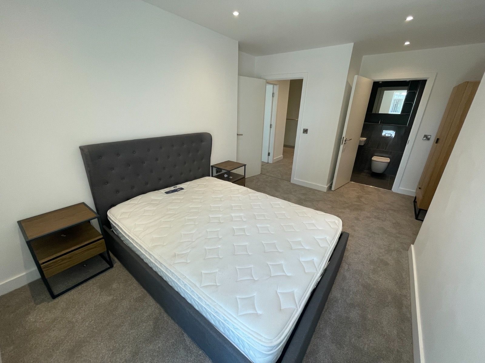 Bedroom with a gray bed, two nightstands, and an open bathroom. Carpeted floor, white walls.