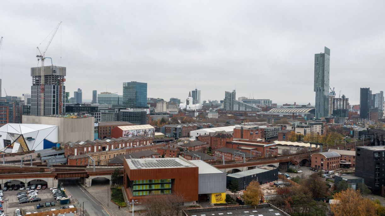 Cityscape of Manchester, UK, under overcast sky. Construction cranes and tall buildings dominate, with older brick structures in foreground.