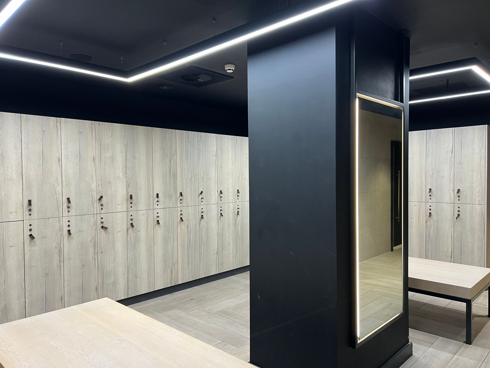 Locker room with wood-look lockers, black pillar, ceiling lights, and benches. Mirror on pillar reflects hallway.