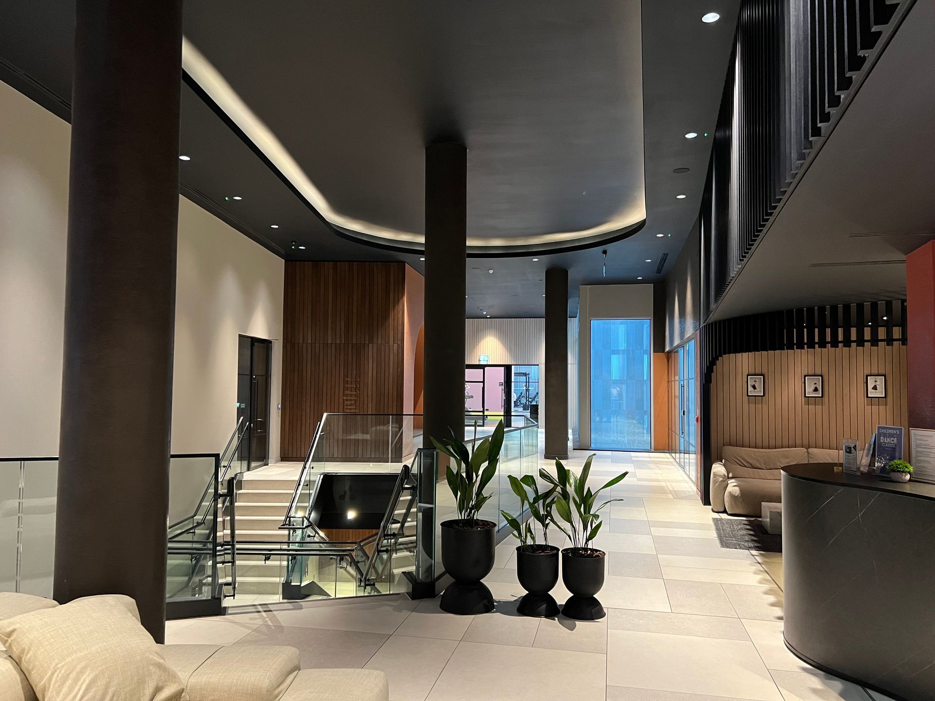 Modern hotel lobby with beige tile floor, dark pillars, and potted plants. Wooden paneling on walls.