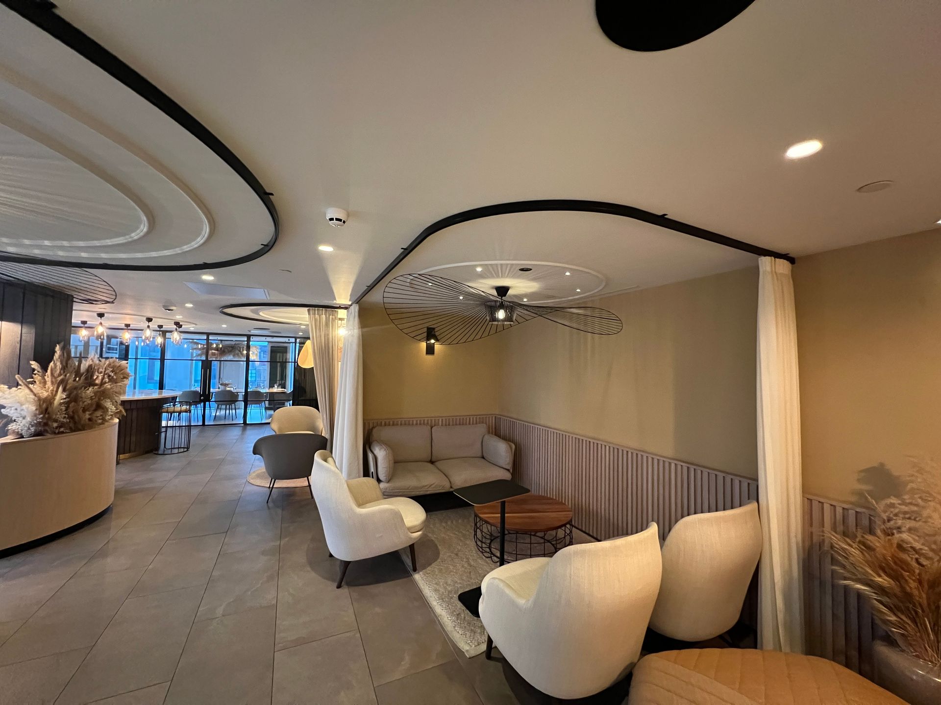 Modern lounge area with curved ceiling, couches, chairs, and small tables. Neutral tones, well-lit with various decor.