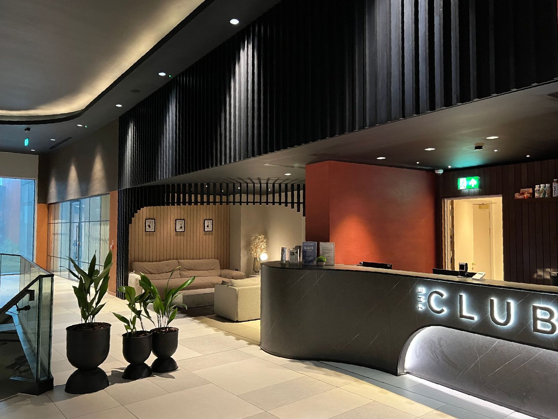 Modern hotel lobby with reception desk, lounge area, and potted plants. Black and wood paneling, neutral tones.