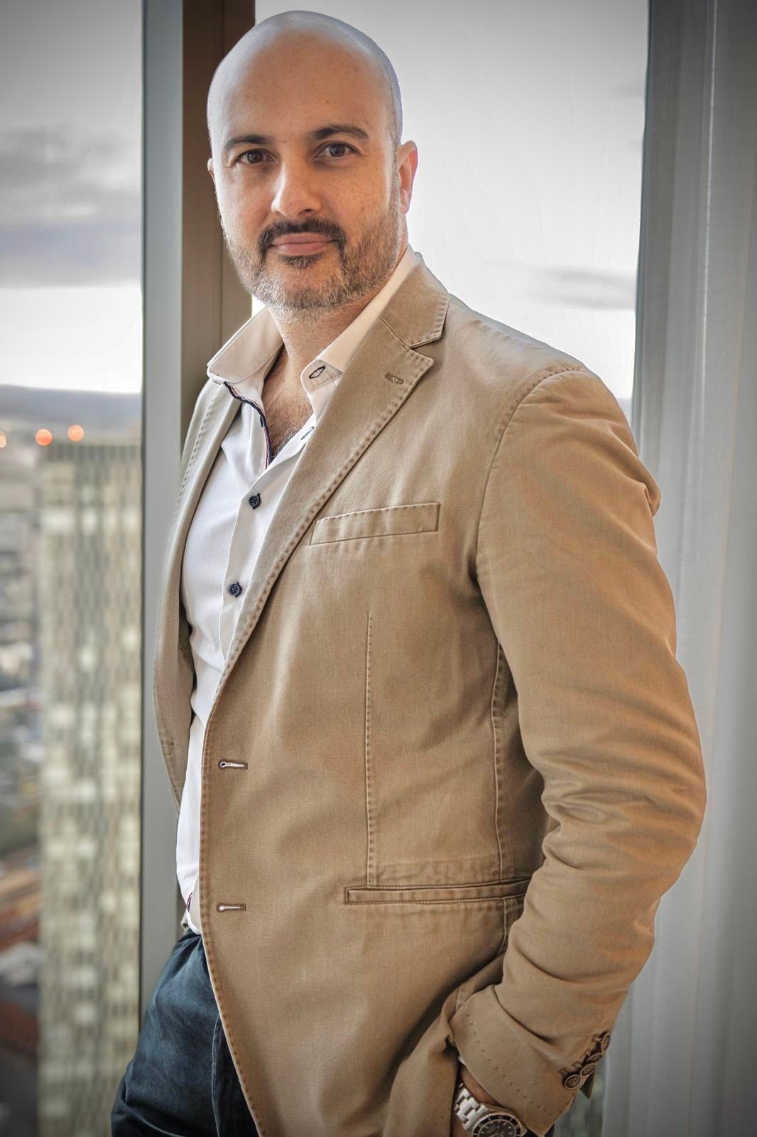 Man in tan blazer, standing by a window, city in background. He looks toward the camera.